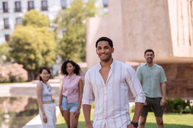 Bir grup arkadaş, bir şehir parkında fotoğraf için poz veriyor. Bir tanesi önde duruyor ve gülümsüyor.