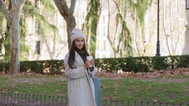 Elinde kahve fincanı olan genç bir kadın, sakin atmosferin ve huzurlu çevrenin tadını çıkararak kış parkında yürüyor.