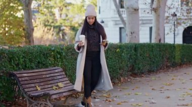 Bej ceketli ve beyaz bereli genç bir kadın bankta oturuyor, kahve içiyor ve akıllı telefonunu kullanıyor.