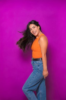 Günlük turuncu bluzlu, kot pantolonlu, saçları dalgalı ve neşeli yüzlü bir kadın.