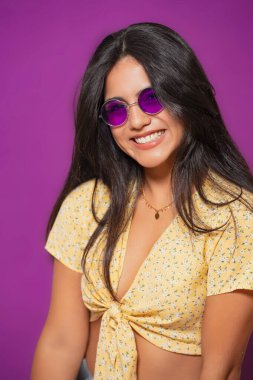 Mor güneş gözlüklü, sarı çiçekli bluzlu, kendine güvenen bir kadın, gülümseyen ve yakın planda parlak mor bir fonda poz veren. 