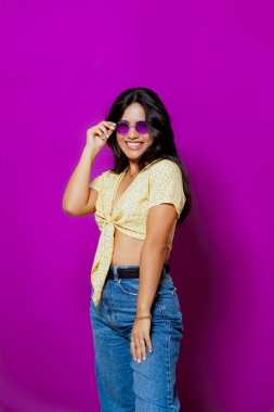 Mor güneş gözlüklü neşeli bir kadın, sarı çiçekli bir bluz ve kot pantolon giyiyor, mor bir arka plana karşı neşeli bir gülümsemeyle güvenle poz veriyor.. 