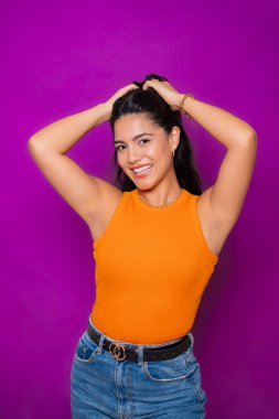 Sıradan turuncu bluzlu ve kot pantolonlu neşeli bir kadın, elleri başının üstünde, parlak bir şekilde gülümsüyor.