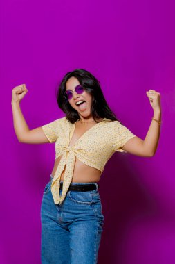 Sarı çiçekli bluz, kot pantolon ve mor güneş gözlüklü kadın, kollarını esnetiyor, canlı mor bir arka plana karşı neşeli bir ifadeyle poz veriyor.. 