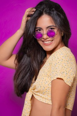Sarı çiçekli bluz ve mor güneş gözlüğü takan şık bir kadın, saçlarını eğlenceli bir şekilde düzeltirken kendine güvenerek gülümsüyor, mor bir arka plana karşı neşeli bir poz veriyor.. 