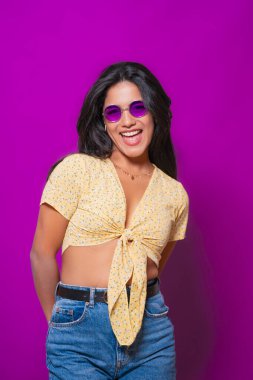 Mor güneş gözlüklü gülümseyen bir kadın ve sarı çiçekli bir bluz, neşeli ve neşeli bir poz veriyor, canlı mor bir arka plana karşı pozitif enerji yayıyor.. 