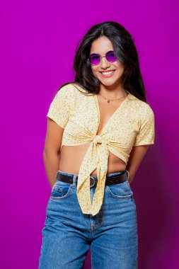 Mor güneş gözlüklü gülümseyen bir kadın, sarı çiçekli bir bluz giyiyor, elleri arkasında canlı mor bir zemin üzerinde güvenle poz veriyor.. 