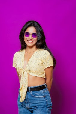 Mor güneş gözlüklü gülümseyen bir kadın, sarı çiçekli bir bluz giyiyor, elleri arkasında canlı mor bir zemin üzerinde güvenle poz veriyor.. 