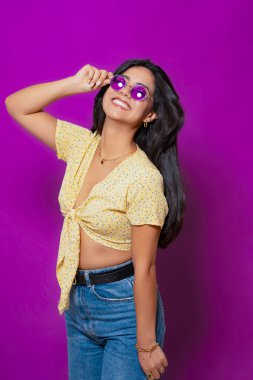 Mor güneş gözlüğü takan neşeli bir kadın ve sarı çiçekli bir bluz, geniş bir gülümseme ve neşeli bir poz. 