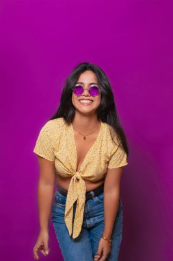 Sarı çiçekli bluz ve mor güneş gözlüğü takan neşeli bir kadın, parlak bir şekilde gülümserken, mor bir arka plana karşı neşeli bir pozda öne eğiliyor.. 