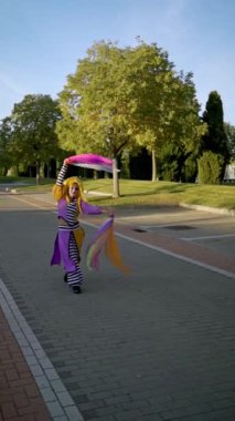 Canlı kostümlü bir sokak sanatçısı güneşli bir parkta ağır çekimde zarif bir şekilde yürürken renkli kurdelelerle hokkabazlık yapıyor.