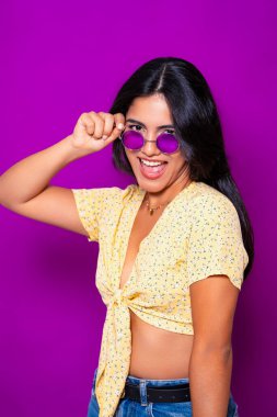 Sarı çiçekli bluz ve mor güneş gözlüklü, neşeli poz veren ve mor arkaplanda gülümseyen şık bir kadın.. 
