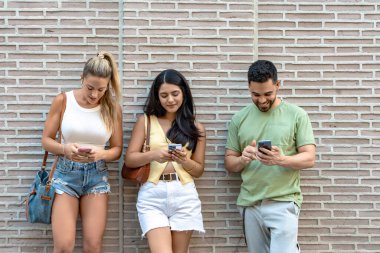 Üç arkadaş duvara yaslanmış telefonlarına bakarken gülümsüyor.