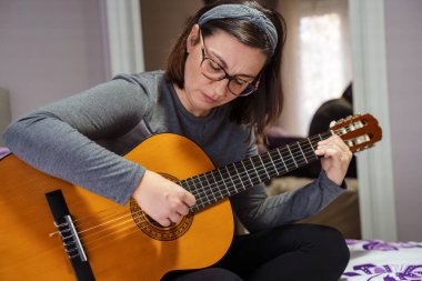Kadın müzisyen akustik gitar çalışırken konsantre oluyor, evde yatakta oturuyor.
