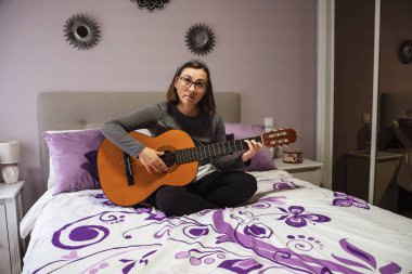 Genç bir kadın, rahat, mor temalı bir yatak odasında yatağında rahat bir şekilde otururken akustik gitar çalışıyor.