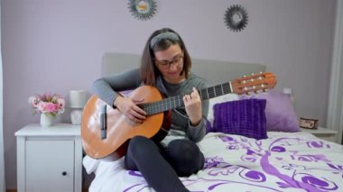 Kadın, bir yatakta oturup, müzik öğrenmenin zorluklarını sergileyerek İspanyol gitarıyla şarkı çalmaya çalışıyor.