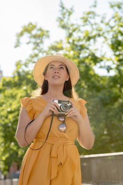 Sarı elbiseli ve güneş şapkalı genç bir kadın dışarıda dikilirken elinde antika bir kamera tutuyor. Güneşli bir günün tadını çıkarırken, tuhaf bir ifadeyle başını kaldırdı.. 