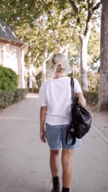 Sırt çantalı genç bir kadın, güneşli bir parkta arkasını kollarken yürüyüp gidiyor.