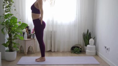 Yoga elbiseli kadın bitkilerle ve doğal ışıkla dolu parlak bir odada bir paspasın üzerine eğiliyor.