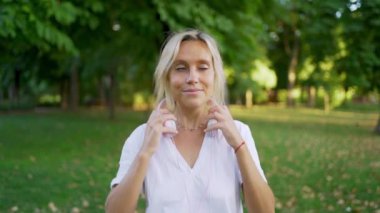 Genç bir kadın, doğa ve yeşilliklerle çevrili güneşli bir parkta kulaklık takarken müzik dinlemekten hoşlanıyor.