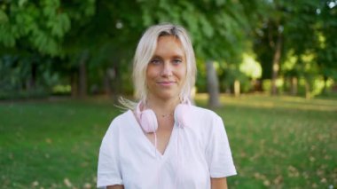 Yavaş çekim montajında kulaklık takarken gülümseyen genç bir kadın güneşli bir parkta müzik dinlemekten hoşlanıyor.