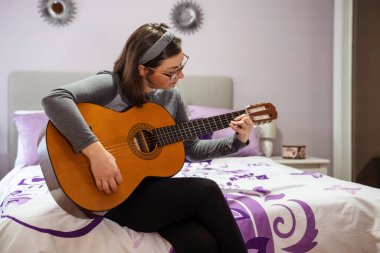 Genç bir kadın akustik gitar çalışıyor, mor bir yatak odasında oturuyor, hobisinin tadını çıkarıyor.
