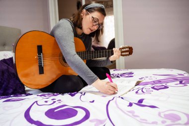 Yeni müzik besteleyen bayan müzisyen akustik gitar çalıyor ve evdeki yatağa notalar yazıyor.