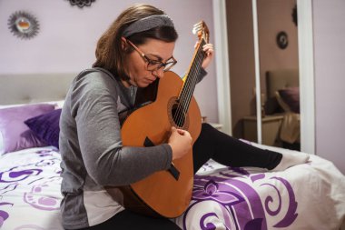 Kadın rahat yatak odasında İspanyol gitarı çalıyor, odayı yatıştırıcı seslerle dolduran ahenkli bir melodi yaratıyor.