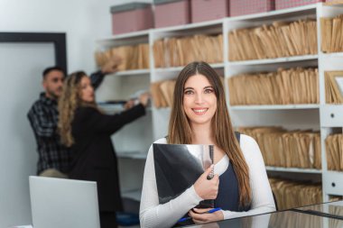 İş arkadaşları arka plandaki dosyaları ararken genç ofis çalışanı gülümsüyor ve elinde belgeler tutuyordu.