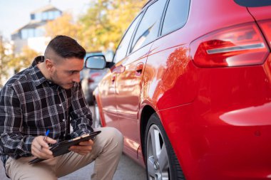 Sigorta acentesi hasarlı kırmızı bir arabanın yanında çömelmiş kaza raporu için not alıyor.