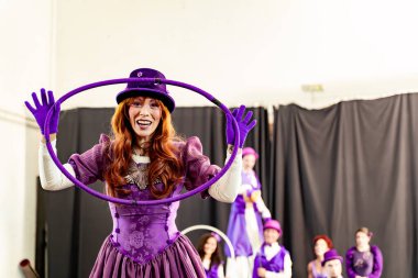 Mor kostümlü kızıl saçlı kadın gülümsüyor ve elinde hula hoop ile arka planda bulanık sirk sanatçıları var.