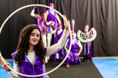 Mor kostümlü genç bir kadın arka planda bulanık sirk sanatçılarıyla birlikte beyaz hula çemberi tutuyor.