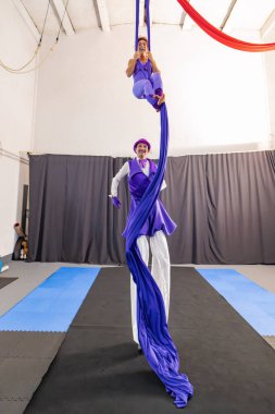 İki sanatçı, hava ipeklerinden sallanan bir kadın ve ayaklık takan bir adam, sirk gösterilerini spor salonunda icra ediyorlar, güç, denge ve sanatçılık sergiliyorlar.