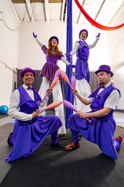 Canlı mor kostümlü dört sirk göstericisi jonglörlüklerini ve stil yürüyüş yeteneklerini bir spor salonunda sergiliyorlar.