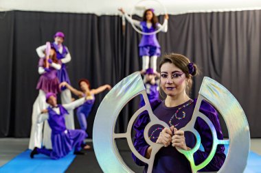 Mor kostümlü sirk göstericileri stil yürüyüşü, akrobatik hareketler ve hula hop dansı yapıyorlar. Ön planda yanardöner bir sahne donanımı var.