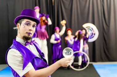 Mor kostüm giyen ve şapka takan sihirbaz sirk sanatçılarıyla kristal küre tutuyor ve arka planda denge kuruyor.