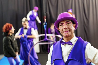 Mor kostümlü ve melon şapkalı gülümseyen sirk sanatçısı arka planda çalışma yapan meslektaşlarıyla sahne arkasında poz veriyor.