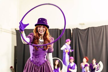 Kırmızı saçlı kadın mor kostüm giyiyor ve hulahop tutuyor, sirk sanatçılarıyla birlikte arka planda, bir gösteri sırasında gülümsüyor