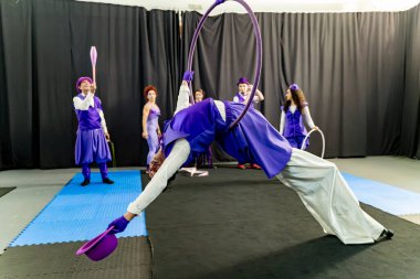 Sirk sanatçısı, diğer sanatçılar izlerken, büyüleyici bir akrobatik gösteri sergileyerek, bir hava çemberinden baş aşağı sarkıyor.