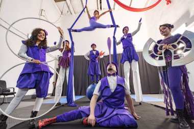 Canlı mor kostümlü sirk sanatçıları hulahop, hokkabazlık ve hava akrobasisi gibi yeteneklerini sergiliyorlar.
