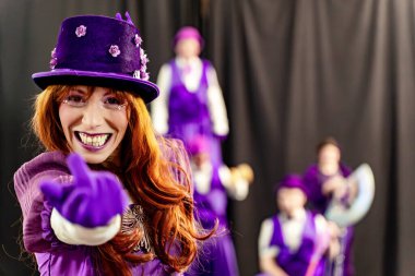 Kırmızı saçlı kadın mor kostümlü, silindir şapkalı, gülümseyen ve işaret eden sirk sanatçıları arka planda dikkatleri dağınık duruyor.