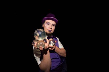 Mor şapka ve yelek giyen sihirbaz, arka planda dört saydam hokkabazlık topunu dengede tutuyor.