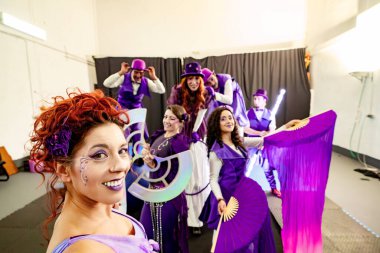 Mor makyajlı kızıl saçlı sirk sanatçısı gülümsüyor ve sahne sanatları grubuyla mor kostümlü poz veriyor.