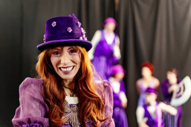 Mor silindir şapka giyen, aynı kıyafeti giyen kızıl saçlı aktris ve sahne arkasında kendine güvenen tiyatro makyajı gülümsemesi.