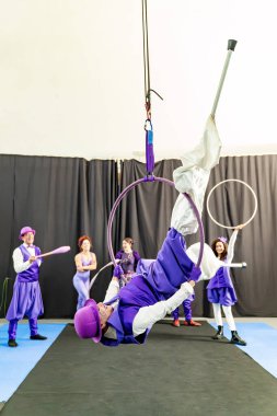 Mor kostüm giyen erkek akrobat, arka planda diğer sirk sanatçılarıyla birlikte baş aşağı asılı olarak hava çemberinde gösteri yapıyor.