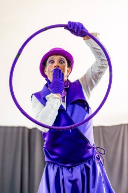 Mor kostüm ve makyaj giymiş pandomimci elinde mor bir hula çemberi tutuyor ve mor eldiveniyle ağzını kapatıyor.