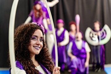 Kıvırcık saçlı, mor makyajlı, gülümseyen ve elinde hula çemberi tutan genç bir kadın.