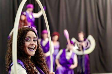 Kıvırcık saçlı ve mor makyajlı genç bir kadın elinde hula hoop tutarken gülümsüyor ve arka planda bulanık sirk sanatçıları var.