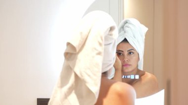 Kafasında havlu olan güzel bir kadın modern bir banyoda aynanın önünde makyaj yapıyor.