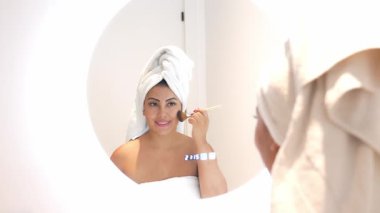 Havlu takan güzel bir kadın duş aldıktan sonra yüzüne fırçayla temeli uyguluyor, aynada kendine bakıyor.